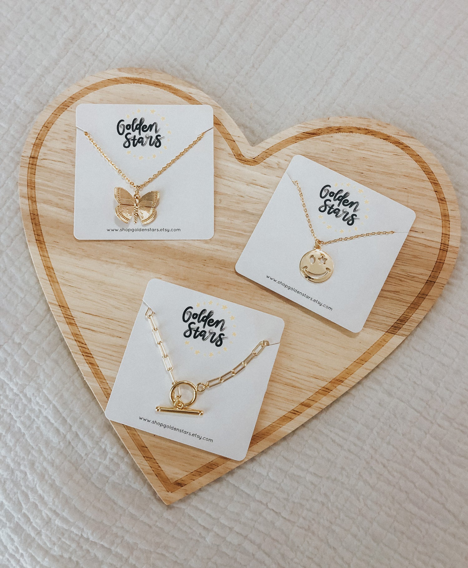 three pieces of jewelry on a wooden block in the shape of a heart. The pieces are our toggle necklace, butterfly necklace, and smiley face necklace.