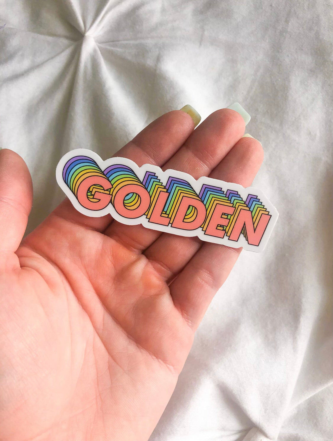 This sticker has the world golden on it in all caps. The sticker is cut so that it indents around where the letters curve. The text is layered with different colors. The first color in the front is pink, then orange, yellow, green, blue and purple.