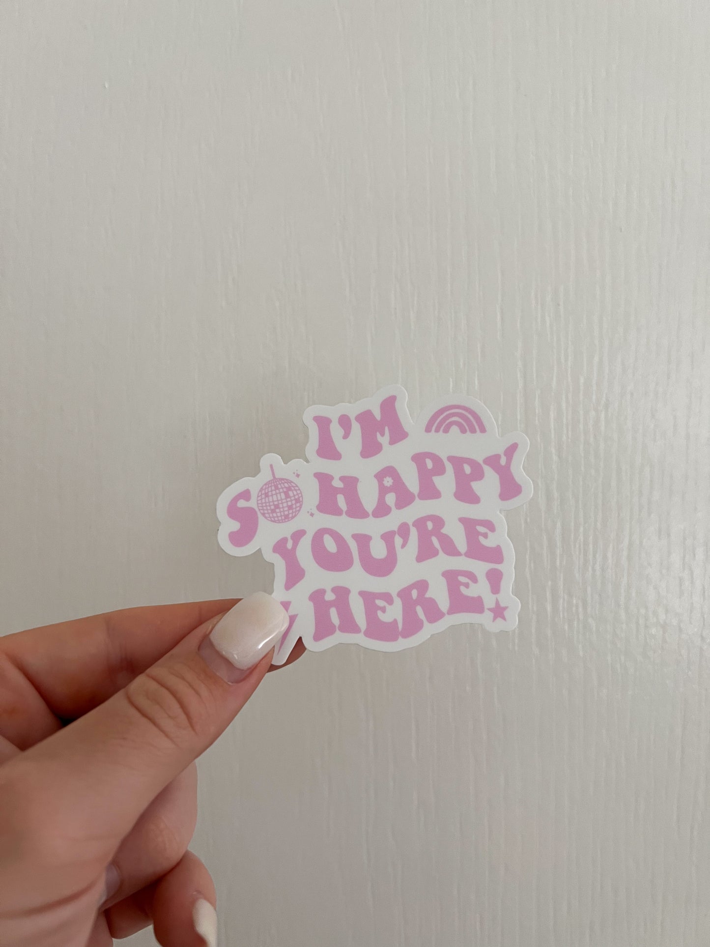 these stickers are a white background with pink letters. The sticker is cutout around the letters so the shape is not a square. It says I'M SO HAPPY YOU'RE HERE! the O in so is a disco ball and there is a lightning bolt before the h in here. the bottom of the exclamation point is a star. there is a rainbow shape (but all pink) after I'M