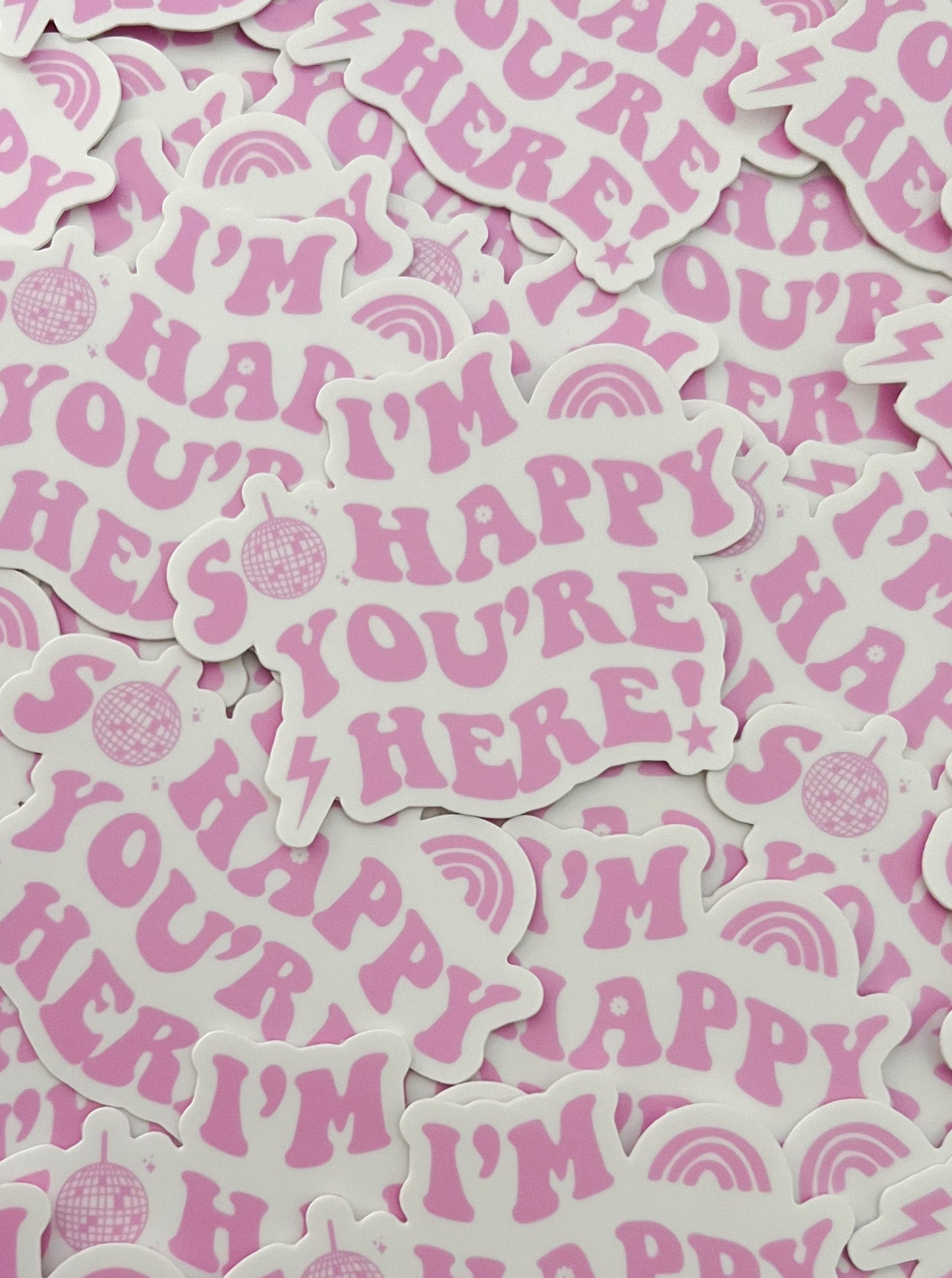 these stickers are a white background with pink letters. The sticker is cutout around the letters so the shape is not a square. It says I'M SO HAPPY YOU'RE HERE! the O in so is a disco ball and there is a lightning bolt before the h in here. the bottom of the exclamation point is a star. there is a rainbow shape (but all pink) after I'M