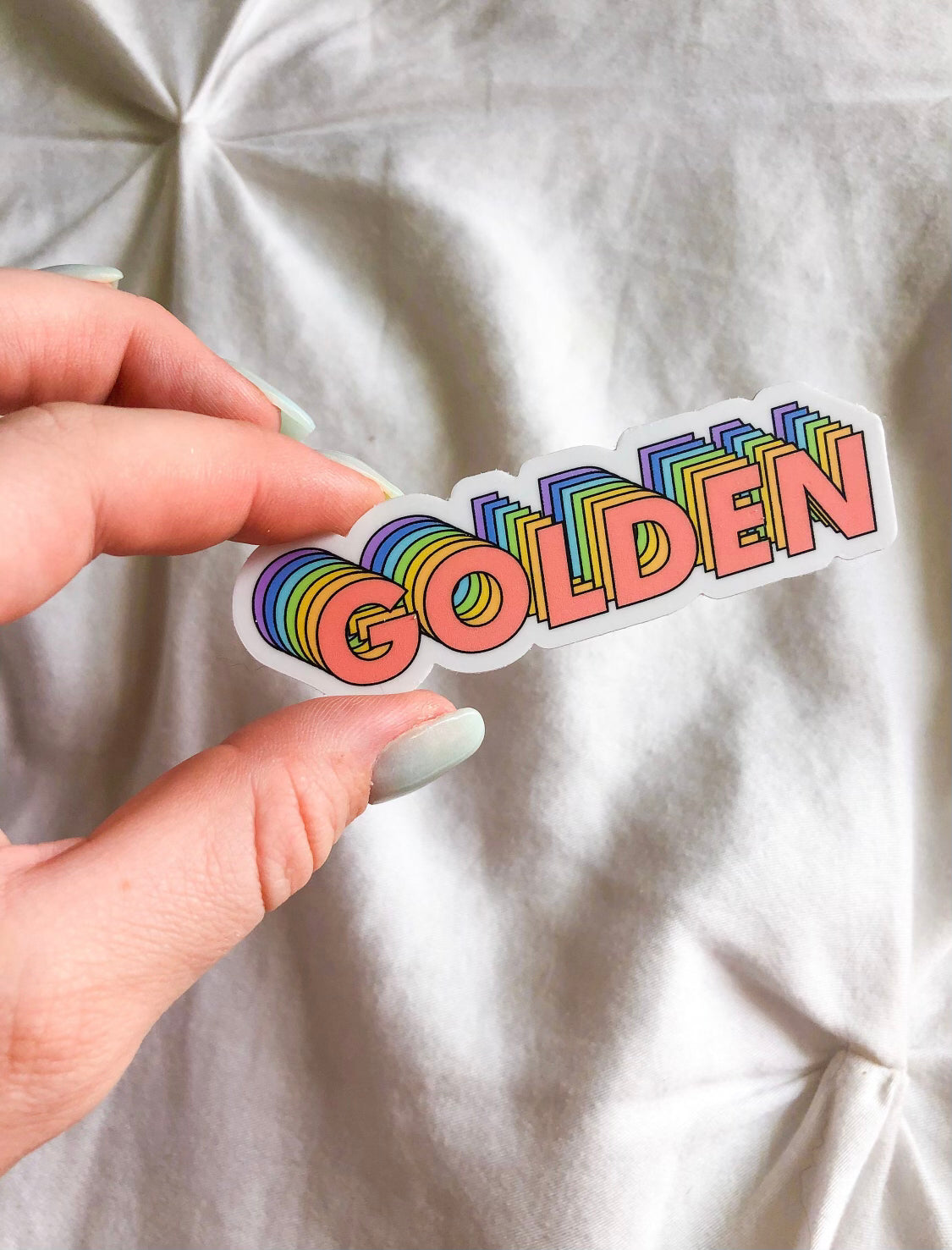 This sticker has the world golden on it in all caps. The sticker is cut so that it indents around where the letters curve. The text is layered with different colors. The first color in the front is pink, then orange, yellow, green, blue and purple.